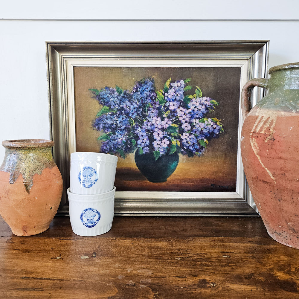 A staged view of a vintage floral oil painting placed on a wooden surface, flanked by rustic earthenware pots, creating a charming and artistic display.