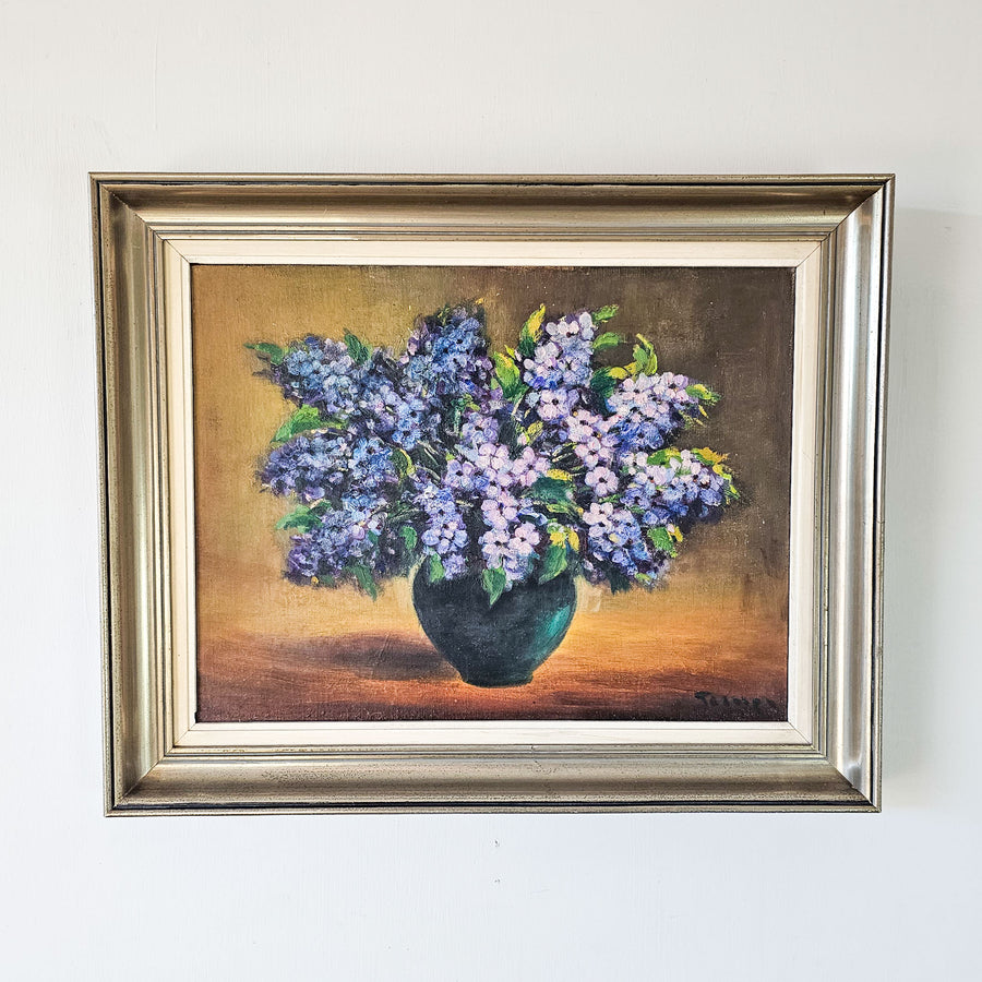Vintage oil painting on board circa 1970 depicting a black vase of small purple flowers set against a warm background. Set within a champagne coloured frame.