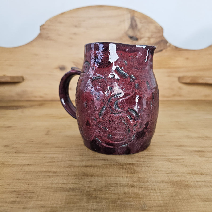 Burgundy glazed studio pottery jug with swirled decorations, blue interior, and sturdy handle. Ideal for serving drinks or displaying flowers. Unique and eye-catching addition to your decor.