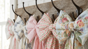 Handmade decorative fabric bows in vintage floral prints hanging from a wooden peg rail against a textured plaster wall.