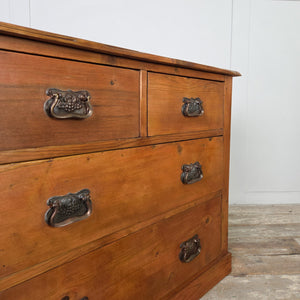 A late 19th to early 20th century farmhouse-style pine chest with four drawers, each fitted with decorative brass handles showing Art Nouveau influence. The warm red-stained and waxed pine finish highlights the grain, and the chest sits on a plain, sturdy plinth base.