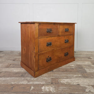A rustic antique pine chest of drawers with a warm red-stained waxed finish. It includes two short drawers above two long drawers, each with decorative brass handles in the Art Nouveau style. The chest rests on a solid plinth base.