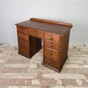 An early 20th century farmhouse-style antique pine desk with nine graduated drawers surrounding a central kneehole. The drawers are finished with original brass knob handles, and the desk is supported by a simple plinth base.
