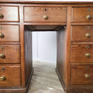 An early 20th century farmhouse antique pine desk featuring nine drawers set around a central kneehole. Each drawer is fitted with an original brass knob handle, and the desk stands on a simple plinth base.
