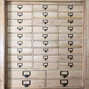 A substantial early 20th-century pine bank of drawers with 34 labelled drawers arranged over a lower cupboard with fixed shelving. The clear wax finish enriches the pine surface, creating a country-style storage unit with extensive capacity for household organisation.