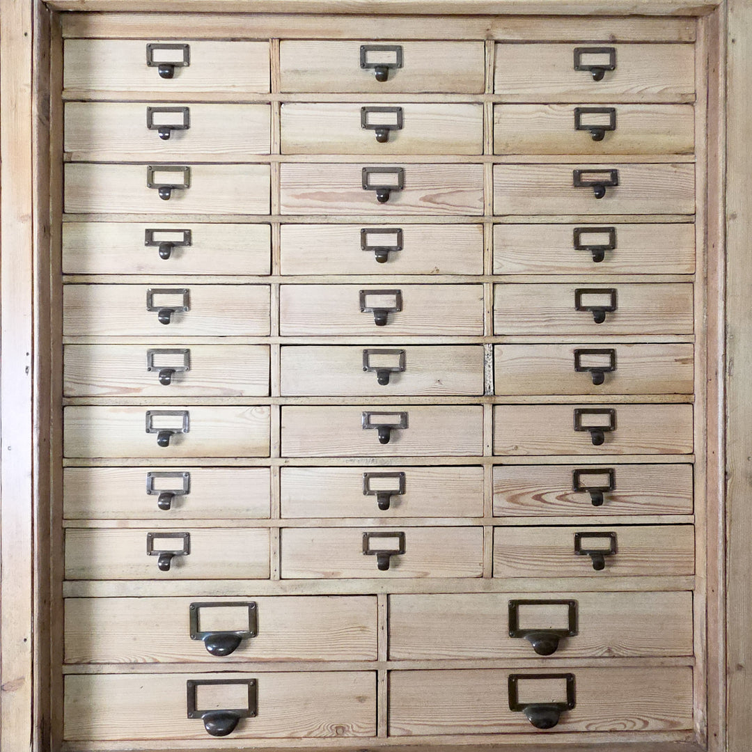 The early 20th century pine bank of drawers combines multiple small brass-handled drawers with a shelved cupboard below, offering extensive and organised storage for home or workplace use.