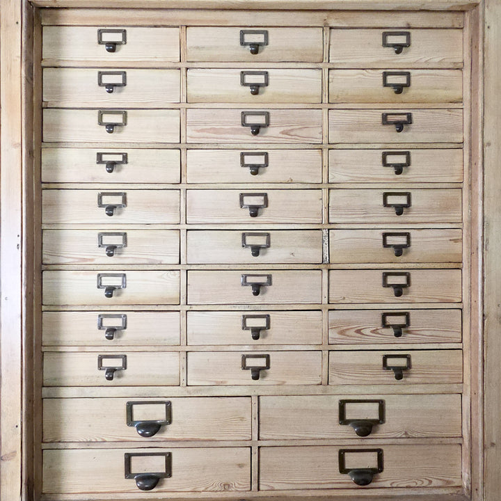 The early 20th century pine bank of drawers combines multiple small brass-handled drawers with a shelved cupboard below, offering extensive and organised storage for home or workplace use.
