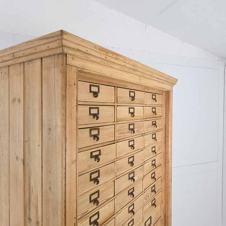 An antique pine bank of drawers with cupboard dating to the early 20th century, with brass label holders on all drawers, and a lower section housing two fixed pine shelves behind panelled doors.