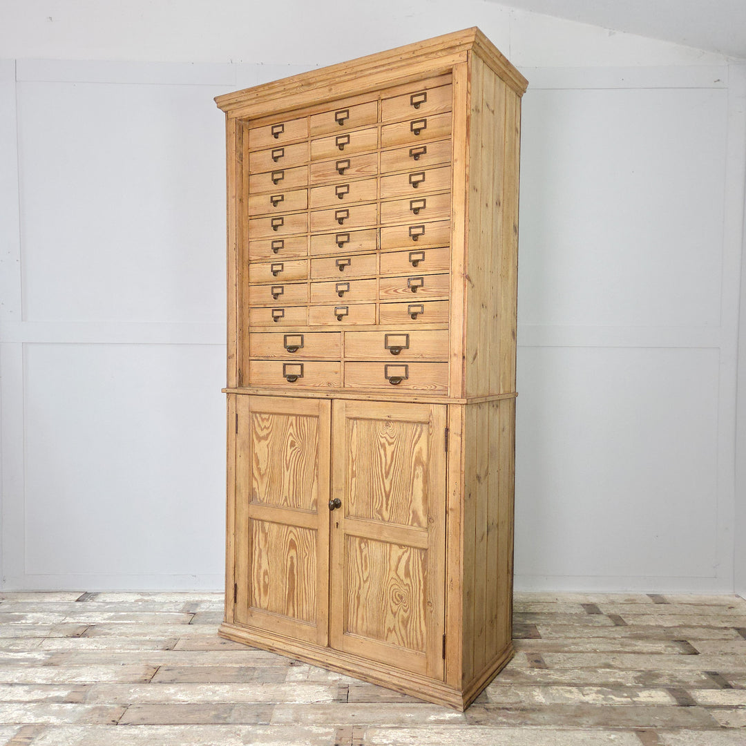 An early 20th century antique pine cabinet with a vertical arrangement of small drawers over a cupboard, each drawer fitted with brass label holders, and two fixed internal shelves for storage.