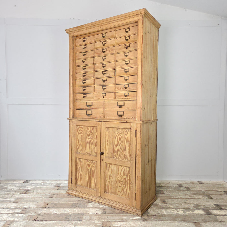 An early 20th century antique pine cabinet with a vertical arrangement of small drawers over a cupboard, each drawer fitted with brass label holders, and two fixed internal shelves for storage.