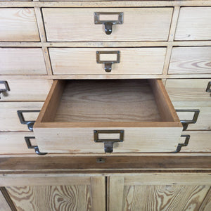 An antique pine bank of drawers from the early 20th century, comprising 34 drawers with brass label holders and a lower cupboard with internal shelves. The clear wax finish enhances the pine grain, creating a practical country-style storage piece with extensive organisation options.