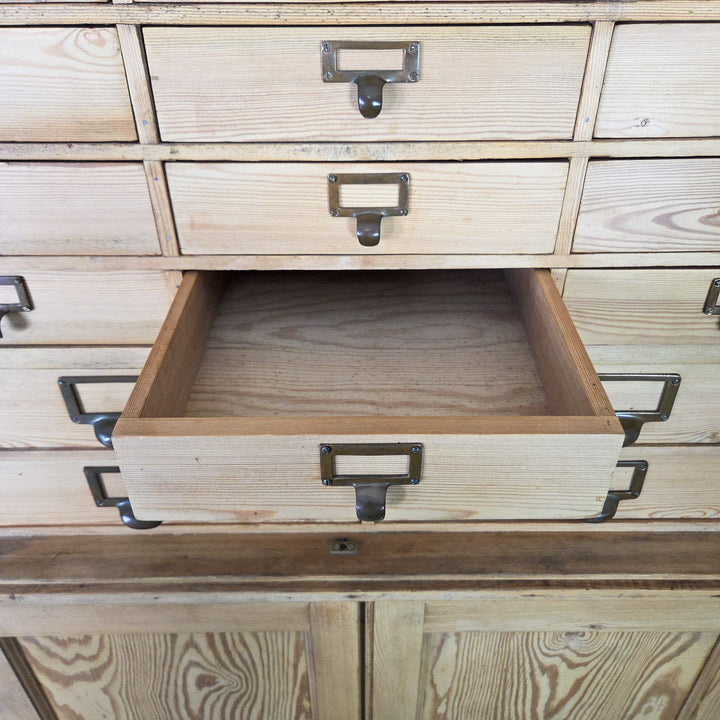 The antique pine bank of drawers from the early 20th century features thirty-four uniform drawers with brass label holders and a lower double cupboard containing two fixed pine shelves.