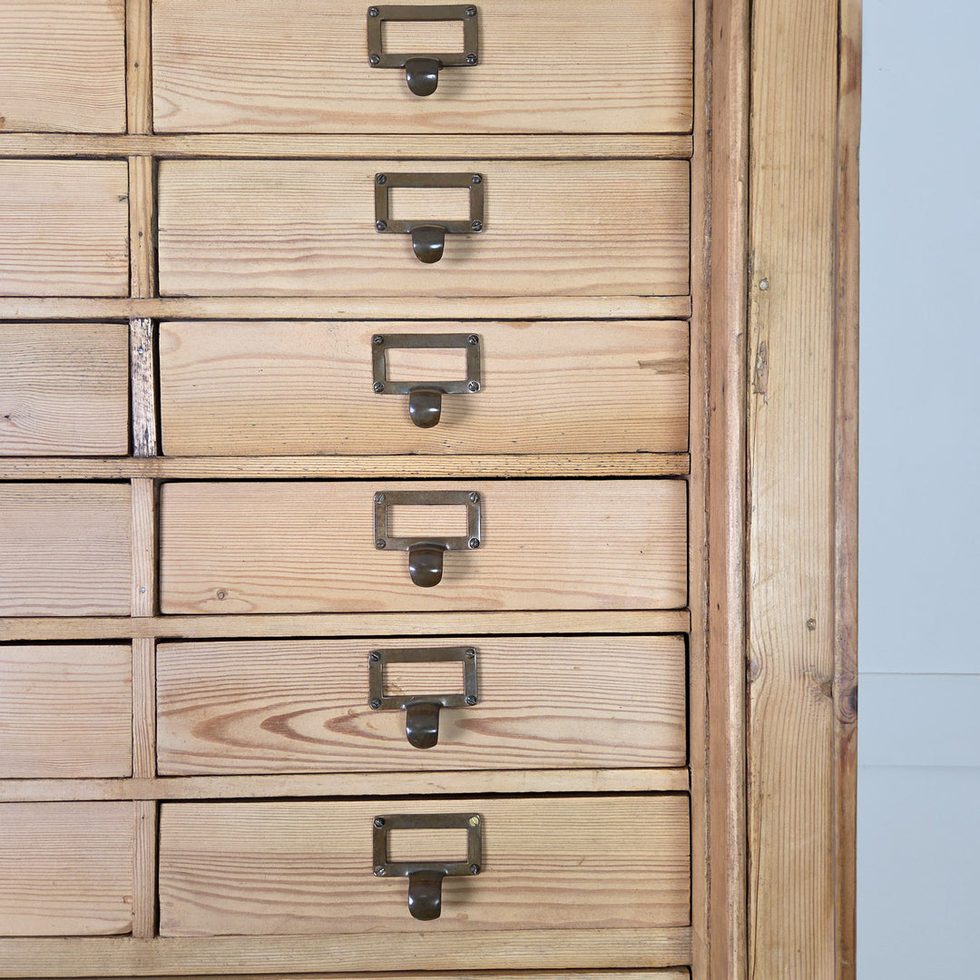 A tall early 20th century pine bank of drawers with brass label holders on each of the thirty-four upper drawers, and a lower panelled-door cupboard with internal shelving, finished in clear wax.