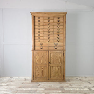 A substantial early 20th century pine bank of drawers featuring thirty-four small brass-handled drawers above a lower cupboard with panelled doors and internal shelving, all in a neutral wax finish.