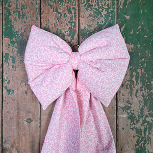 A farmhouse-inspired handmade bow made from genuine vintage Laura Ashley “Firenze” cotton in a soft pink damask design. Structured with padding and tapering tails, finished with an antiqued brass hoop. Works as a hanging bow, door bow or country decorative accent.