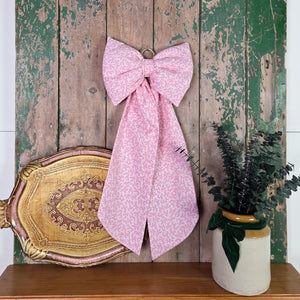 A structured handmade bow created from vintage 1985 Laura Ashley “Firenze” cotton, featuring a delicate pink damask flourish print. This farmhouse-style hanging bow includes padded shaping, tapering tails and a brass hoop, suitable as a decorative door bow or cottage-style accent.