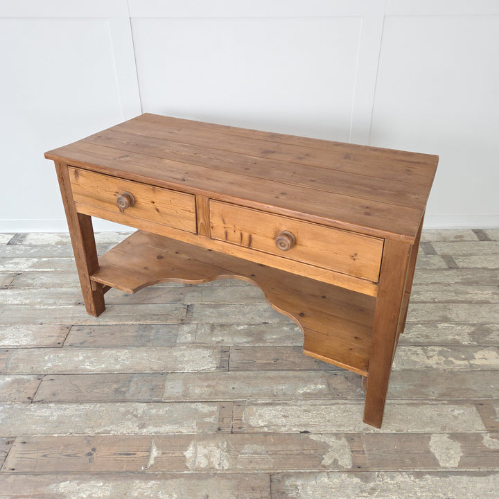 A Victorian antique pine farmhouse prep table dating to the 19th century with a broad planked top, two drawers with original knobs and a shaped lower shelf. Raised on chunky straight legs, this antique pine table can be styled as a rustic kitchen prep surface, a hallway console, a serving table in a dining space or adapted as a farmhouse sideboard or work table for storage and display.