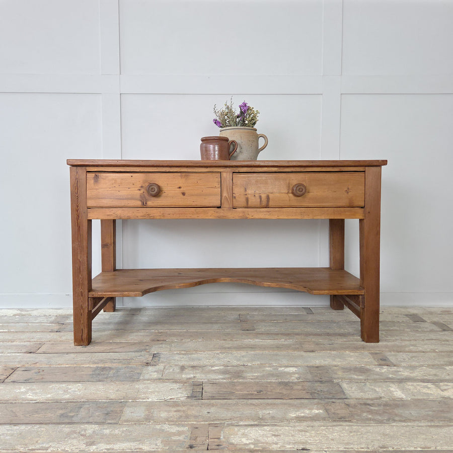 A Victorian antique pine farmhouse prep table from the 19th century with a solid planked top, two drawers fitted with original wooden knobs and a shaped lower shelf for storage or display. Raised on chunky straight legs, this versatile antique pine table can be used in a kitchen as a prep or serving table, in a hallway as a rustic console, or adapted as a sideboard or work table within a farmhouse or country interior.