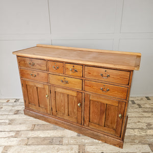 A cottage style 19th-century pine dresser base with seven drawers in assorted sizes, three cupboards and brass swan-neck handles. The lighter scrubbed pine top contrasts with the darker base, offering generous storage throughout the home.