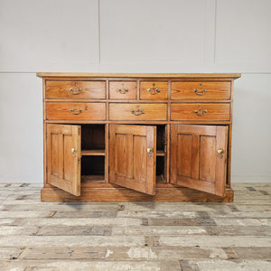 A rustic pine farmhouse style dresser base with seven drawers, three cupboards and matching brass hardware. The scrubbed pine top, lighter in tone, sits above the deeper 19th-century base, forming a practical sideboard.