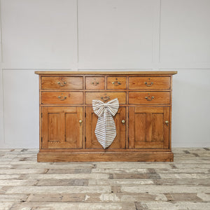 A farmhouse style 19th-century pine dresser base featuring seven assorted drawers and three lower cupboards, all with matching brass swan-neck handles and round knobs. The scrubbed pine top with cleated ends completes this substantial sideboard for home storage.