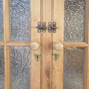 A large antique pine cupboard dating from the early 20th century, featuring glazed double doors with frosted glass and adjustable internal shelving. Finished with a waxed finish, this country cupboard offers generous storage and retains a straightforward, practical rustic character.