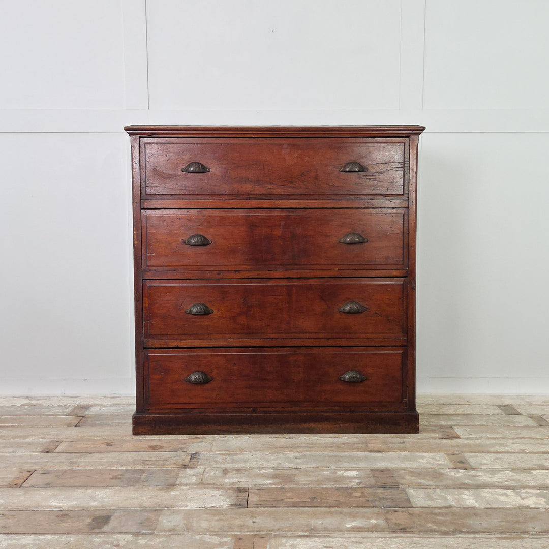 A substantial antique pine chest dating from the early 20th century, featuring four well-proportioned drawers fitted with brass cup handles. Made from solid pine with a dark stained finish and panelled sides, raised on a plinth base with visible wear that reflects its age.
