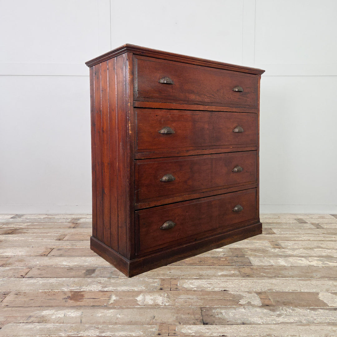 A four-drawer antique pine chest from the early 1900s, finished in a dark stain that enhances the aged surface. Each drawer is fitted with brass cup handles, the sides are tongue-and-groove panelled, and the piece stands on a simple plinth base with solid construction.