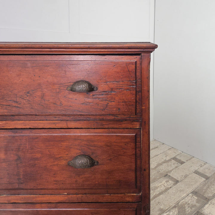An early 20th-century pine chest featuring four drawers with brass cup handles and a dark stained surface. The simple moulded top, panelled sides, and plinth base highlight the sturdy proportions and practical design of traditional country furniture.
