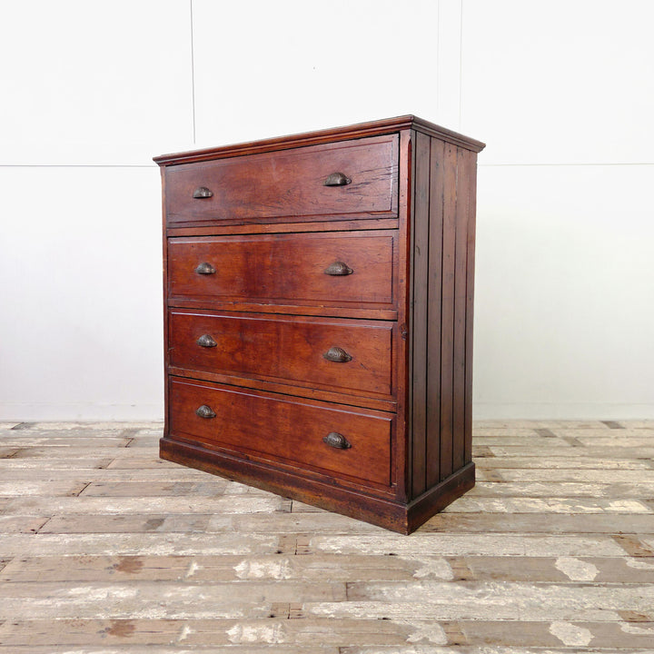 An early 20th-century antique pine chest of drawers with four full-width drawers and decorative brass cup handles. Constructed from solid pine and finished in a rich dark stain, the piece stands on a simple plinth base with tongue-and-groove panelled sides showing gentle age and use.
