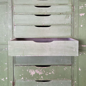 An antique French green painted cupboard featuring two doors and eight interior drawers with slatted bases, arranged in a graduated layout. The shaped plinth and projecting cornice lend a country farmhouse feel, while the original paint highlights the age and character of this practical storage cupboard.