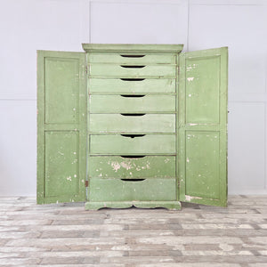 An antique French cupboard from the early 20th century in its original soft green paint, with two panelled doors opening to eight drawers and slatted bases. The shaped plinth and cornice give this wooden painted cupboard a country house feel, offering practical and organised internal storage.