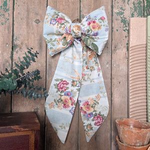 A large handmade decorative fabric bow created from vintage floral stripe fabric, part of the Jennings & Cox Country House collection. With a padded form, long tapering tails, and brass hanging ring, this piece is perfect for doors, mantels, or as a thoughtful gift.