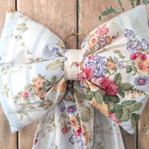 Handmade decorative Country House Bow crafted from vintage floral stripe fabric in a soft blue and white design with colourful flowers. Featuring a padded body, tapering tails, and a brass ring, this bow is ideal for rustic interiors, weddings, or seasonal styling.