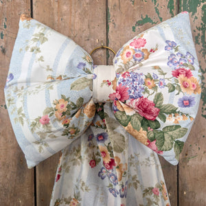A large handmade decorative fabric bow crafted from vintage floral stripe material in soft tones, featuring a padded form and long tails. This Jennings & Cox Country House Bow is completed with a brass ring for hanging and is designed to bring vintage character to doors, mantels, or interiors.