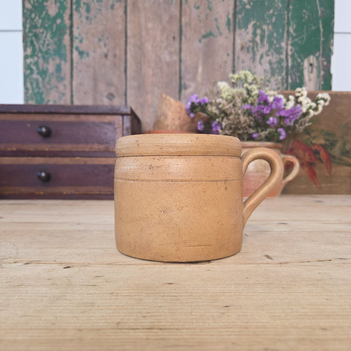 Vintage French stoneware confit pot with single handle and glazed surface in neutral tones. Crafted from durable earthenware, this traditional French kitchen pot was once widely used for food preparation and storage. It remains functional today, suitable for utensil storage or displaying small plants such as herbs or succulents.
