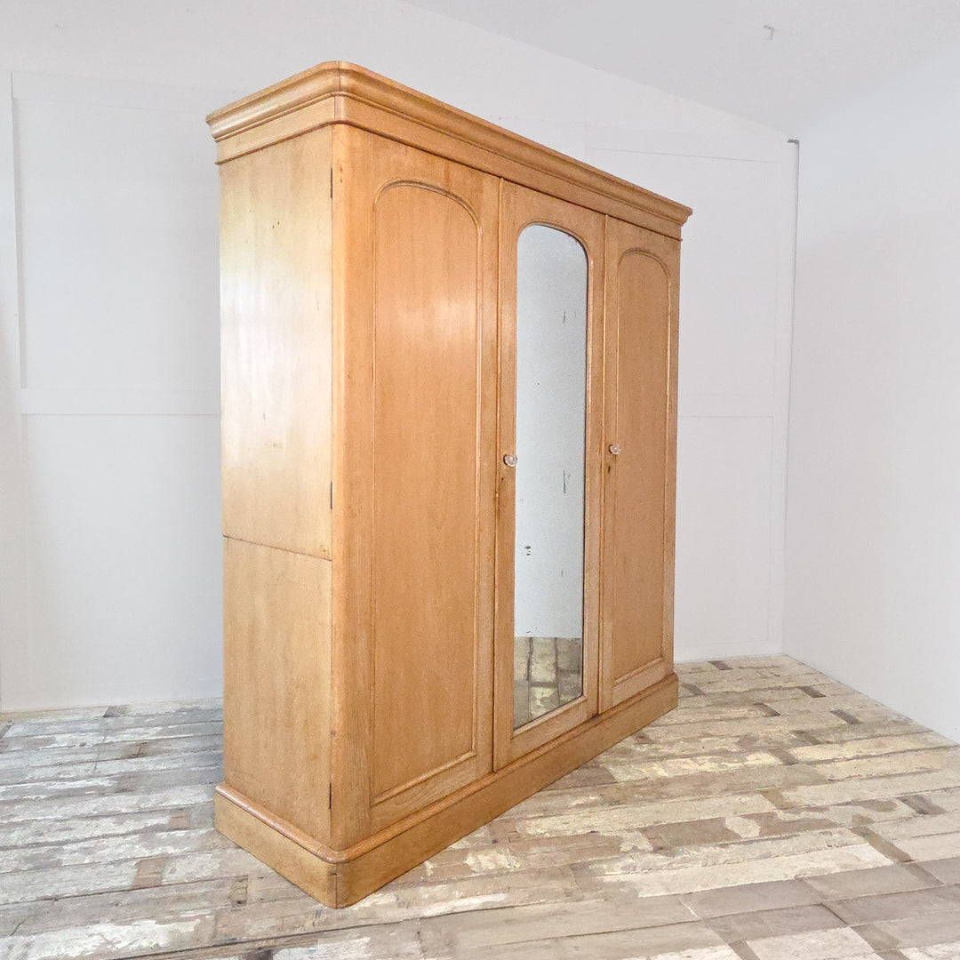 Antique pine triple wardrobe from the Victorian period with mirrored centre door, brass fittings, and solid pine construction. The wardrobe features hanging rails, drawers, and a lift-up compartment, offering practical 19th century bedroom storage in authentic antique pine.