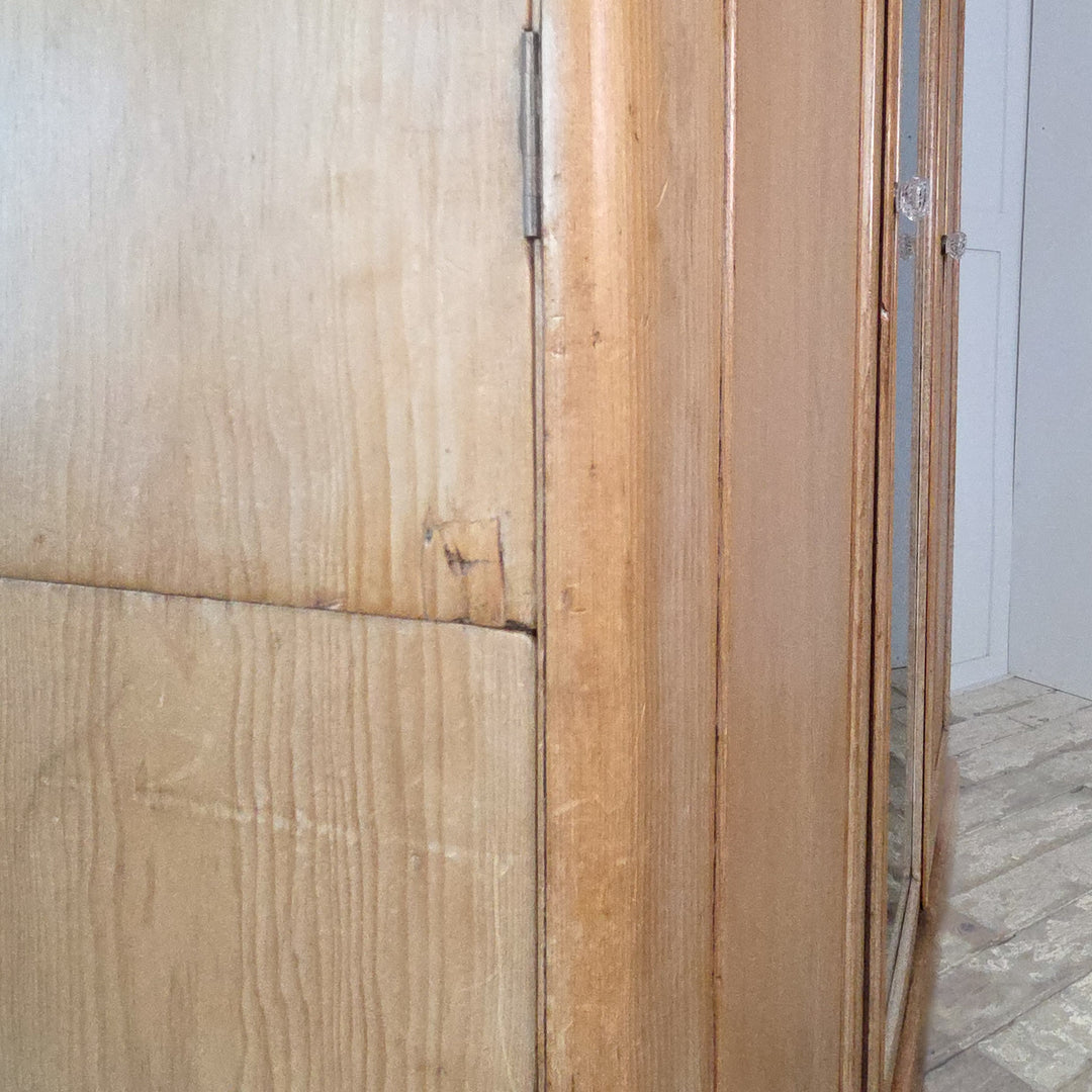Victorian triple wardrobe crafted in antique pine, featuring a mirrored centre door and two panelled doors. The left side contains four drawers beneath a hanging rail, while the right side offers further hanging space, a tray, and a hidden storage area. A well-built 19th century pine wardrobe typical of Victorian furniture.