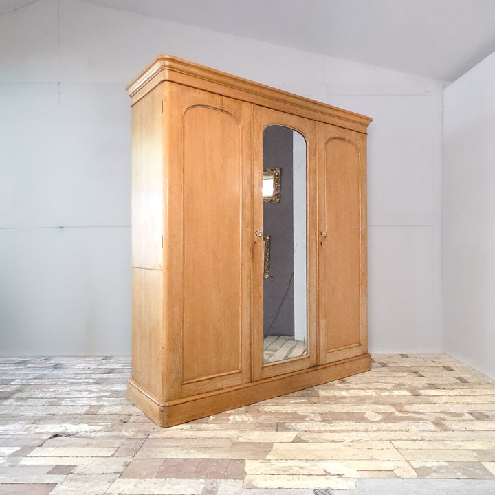 A substantial 19th century Victorian pine wardrobe with triple doors, mirrored centre, and internal storage configuration. This antique pine wardrobe combines drawers, hanging rails, and compartments, reflecting the durable construction and understated design of Victorian pine furniture.