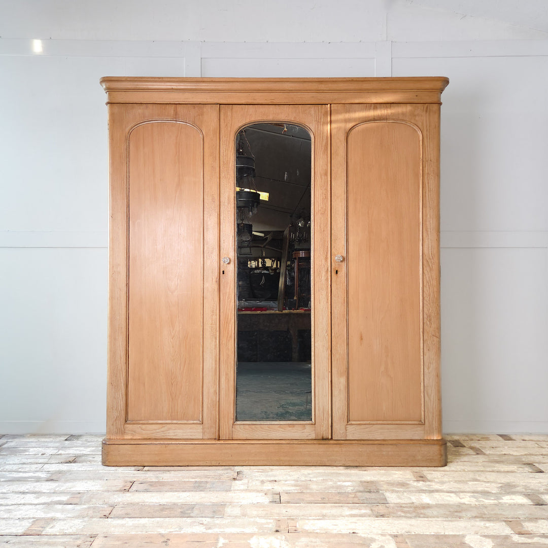 A large antique Victorian pine triple wardrobe from the late 19th century, featuring a mirrored central door and two outer panelled doors. This 19th century wardrobe combines drawers, hanging rails, and a hidden compartment, making it an excellent antique pine storage piece. A substantial example of Victorian bedroom furniture crafted from solid pine with brass swan-neck handles and glass door knobs.