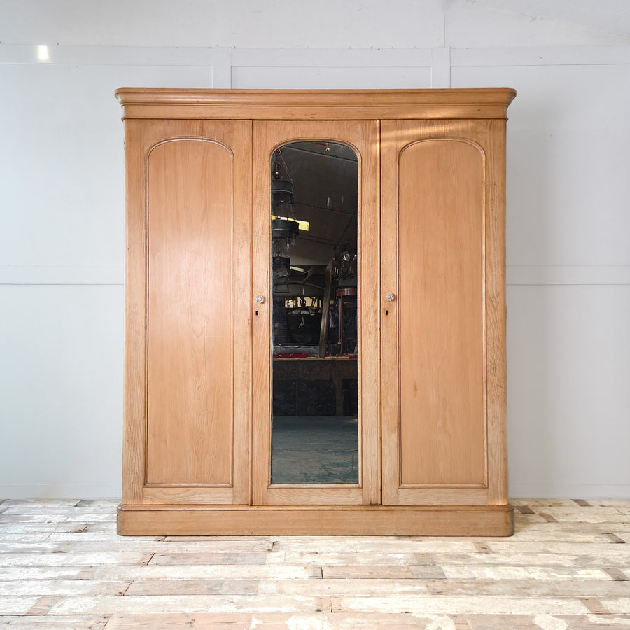 A large antique Victorian pine triple wardrobe from the late 19th century, featuring a mirrored central door and two outer panelled doors. This 19th century wardrobe combines drawers, hanging rails, and a hidden compartment, making it an excellent antique pine storage piece. A substantial example of Victorian bedroom furniture crafted from solid pine with brass swan-neck handles and glass door knobs.