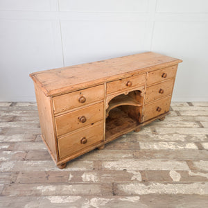 An antique pine dresser base from the Victorian period with a central open shelving section and seven drawers of graduating depth. The drawers retain their original wooden knobs, making it suitable for use in a kitchen or hallway setting.