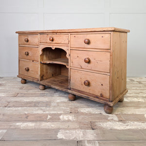An antique Victorian pine dresser base featuring seven drawers flanking a central open shelving section. Raised on bun feet with a moulded top edge, it can be used as a sideboard in a living space or for storage in a hallway.