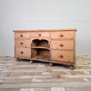 An antique pine dresser base dating from the Victorian period, with seven drawers arranged around a central open shelving section. The piece stands on bun feet and works well in a kitchen or hallway for storing everyday household items.