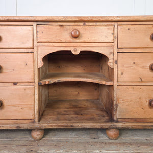 An antique pine dresser base from the Victorian era with seven drawers and a central open shelving section. The drawers and shelves make it suitable for use in a kitchen or as storage in a hallway.