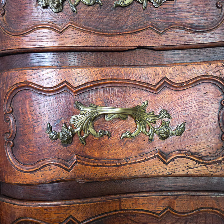 A 19th century French walnut serpentine chest of drawers with four carved drawers, ornate brass handles, and escutcheons. The walnut surface has aged to a warm patina, and the chest stands on short carved legs, maintaining its strong form.