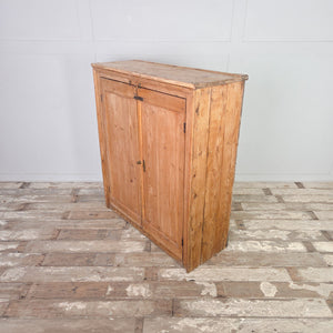 Victorian pine cupboard with panelled double doors, brass hardware including a knob and locking bolt, and a simple plinth base beneath a shelved interior.
