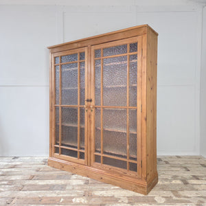 An antique pine bookcase with frosted glazed doors, solid pine frame, and natural waxed finish. Inside, four adjustable shelves provide useful storage for books, tableware, or linens.