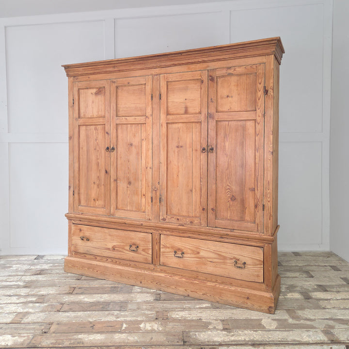 An antique Victorian English pine housekeeper’s cupboard featuring four panelled doors to the top section, fixed shelves inside, and two deep base drawers below, all supported on a plinth base with cornice detailing.