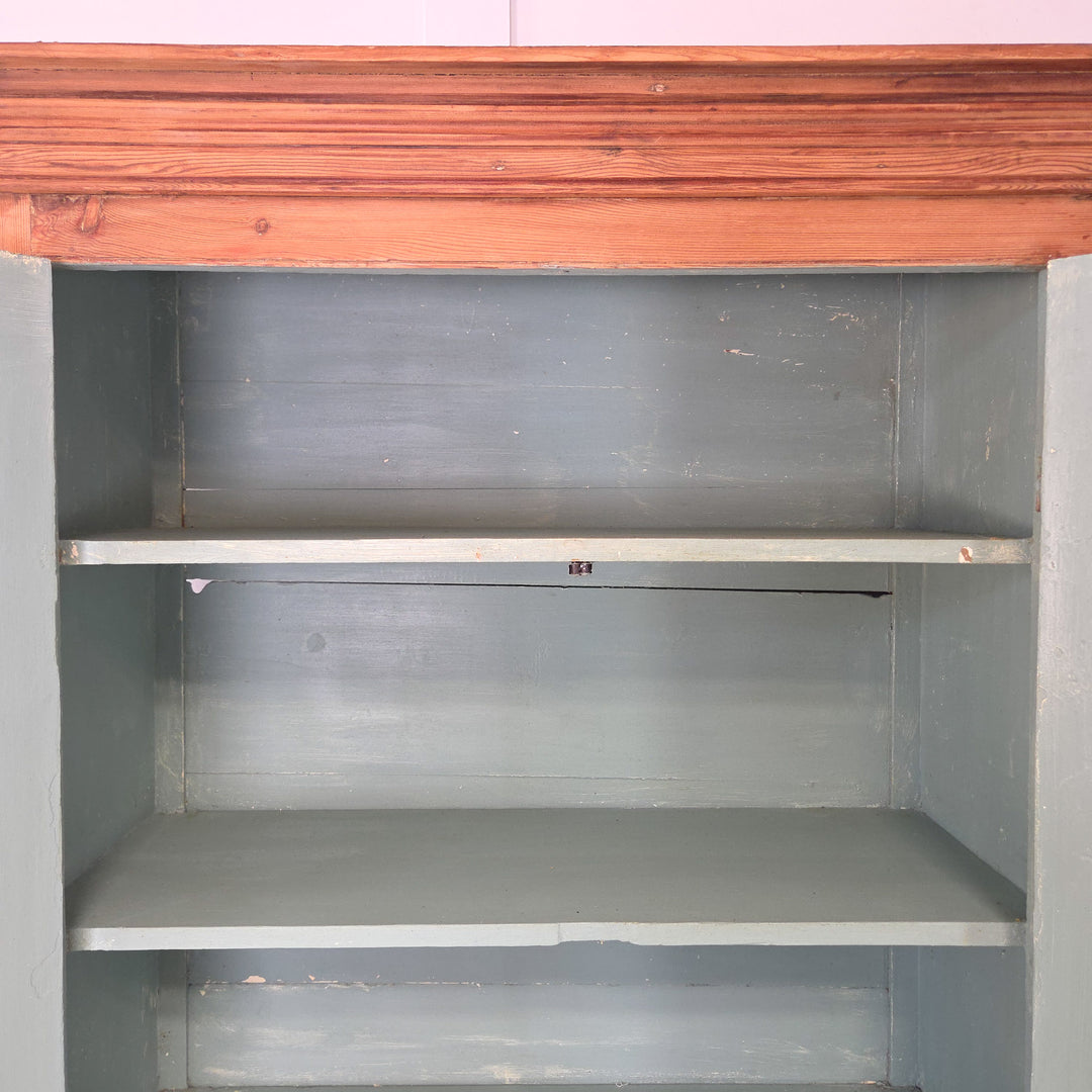 • The antique Victorian pine housekeeper’s cupboard has a substantial pine frame, upper panelled cupboard doors opening to blue-painted shelving, two lower drawers with brass handles, and a stable plinth base.
